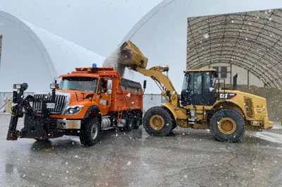 A vehicle loads salt into a salt truck with snow flying.